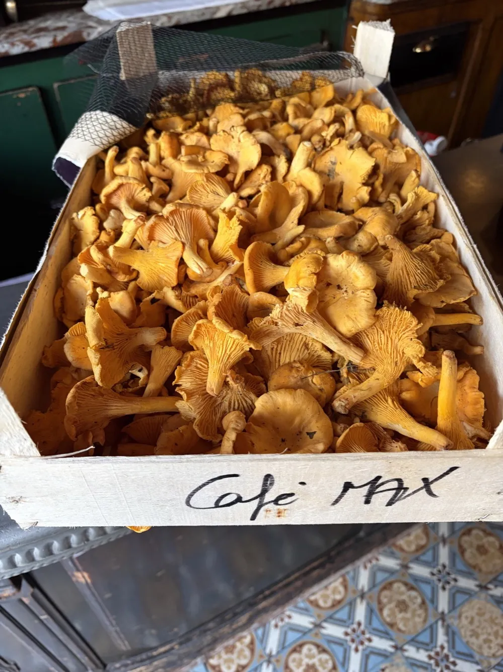 Girolles de Sologne fraîches dans une cagette en bois marquée Café Max, au-dessus du carrelage ancien du bistrot