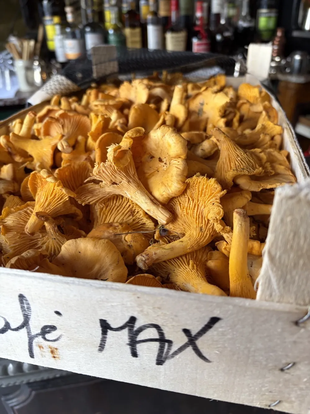 Cagette de girolles de Sologne marquée Café Max au feutre, posée sur le comptoir devant les bouteilles du bar